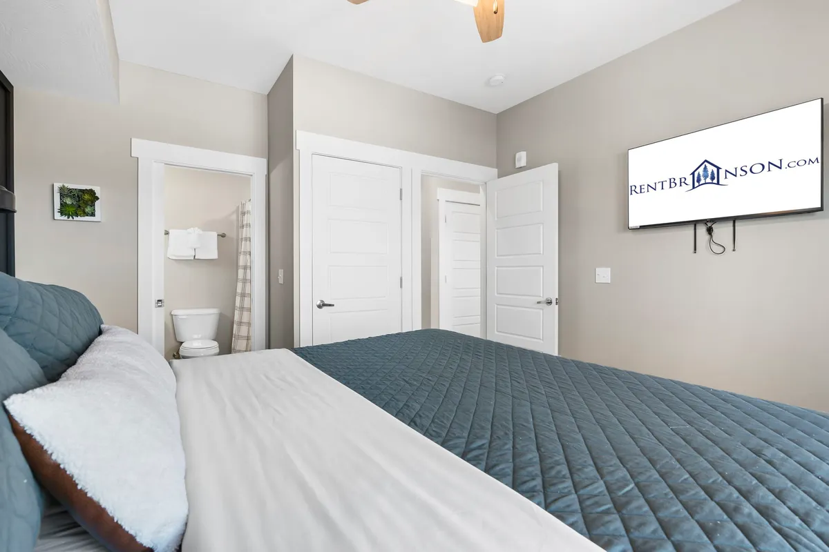 A king or queen suite bedroom featuring a comfortable bed dressed in a blue quilt, a wall-mounted television, and an open door leading to a private bathroom. The space is modern, cozy, and well-lit, with soothing colors and soft bedding.