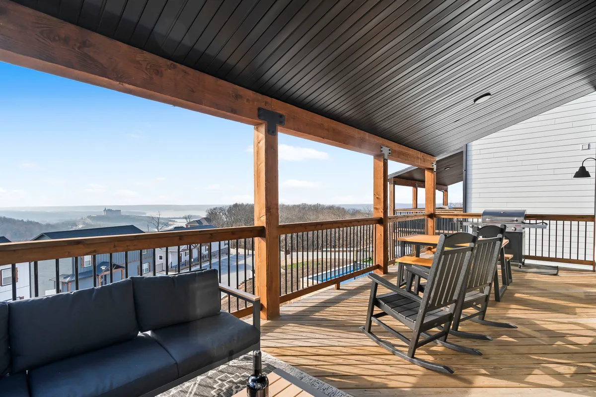A deck featuring plush outdoor seating, a wooden railing, and a scenic view of the surrounding landscape. The area is furnished for comfort and socialization, with rocking chairs, an outdoor sofa, and a grill for entertaining.
