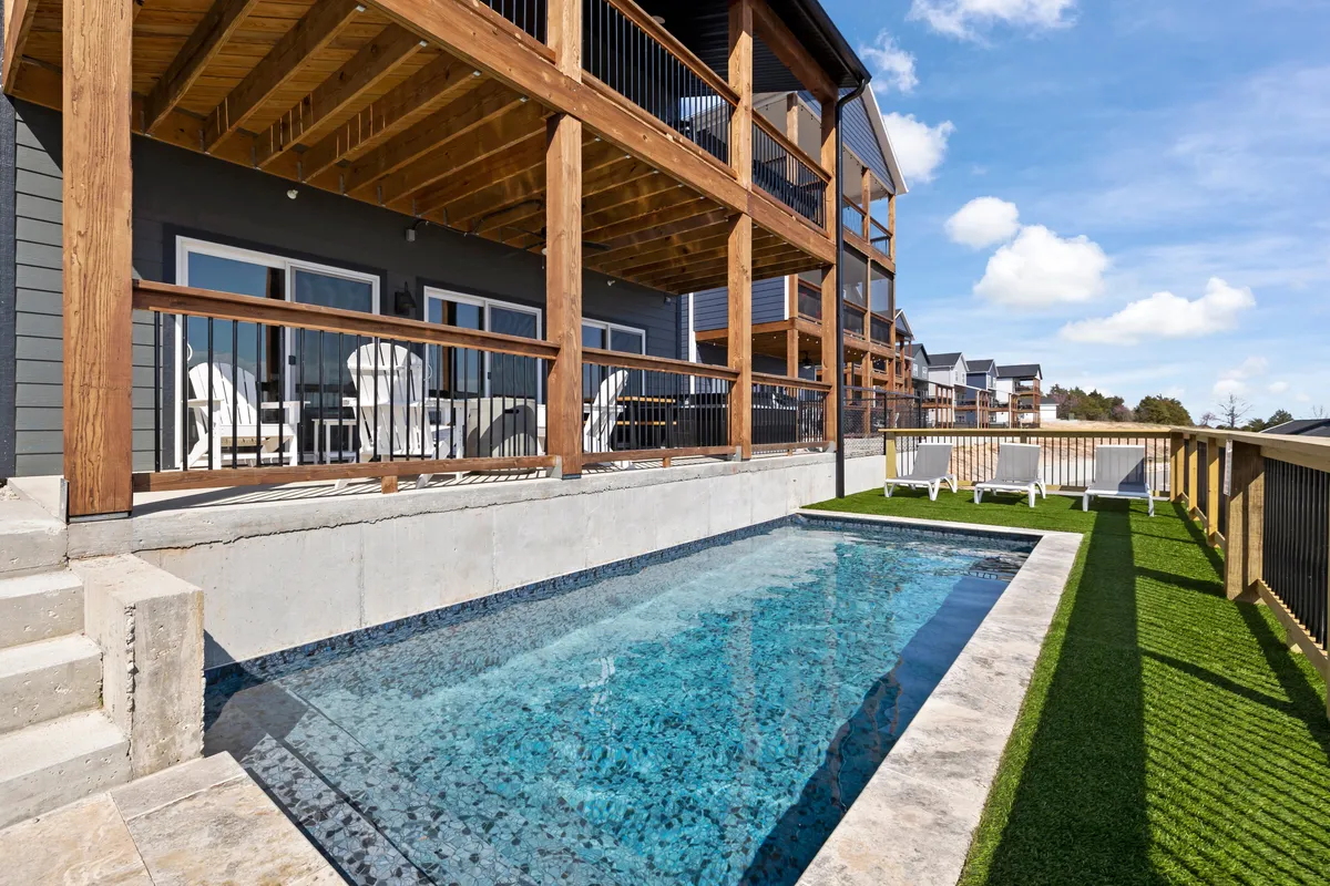 The image showcases the exterior of Rocky Top Lodge at Chateau North, featuring a sparkling private swimming pool surrounded by a wooden deck with Adirondack chairs, set against a lush, green lawn. The scene is perfect for swimming, sunbathing, relaxing, or gathering with friends and family, with hints of sunset lighting that evoke warmth and tranquility.