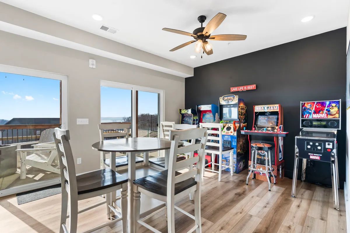 This image presents a vibrant lower-level space featuring arcade machines, a spacious dining table, large windows, and a ceiling fan. The visible arcade machines and table setup suggest an environment perfect for energetic gatherings, friendly competitions, and family game nights, capturing the essence of a private arcade and game lounge.