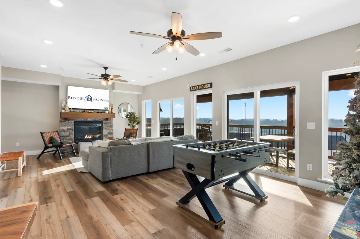 A cozy game lounge featuring a foosball table, comfortable seating, a fireplace, and expansive windows with views of the lake. The space is designed for relaxation as well as entertainment, blending comfort with fun and offering a glimpse outdoors, hinting at patio access.